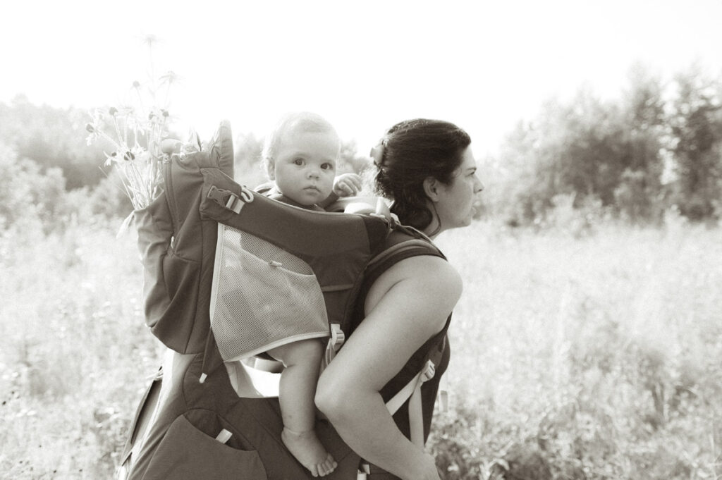 Baby in a backpack carrier on mom's back with flowers in a Sudbury field documented by a Sudbury photographer. 